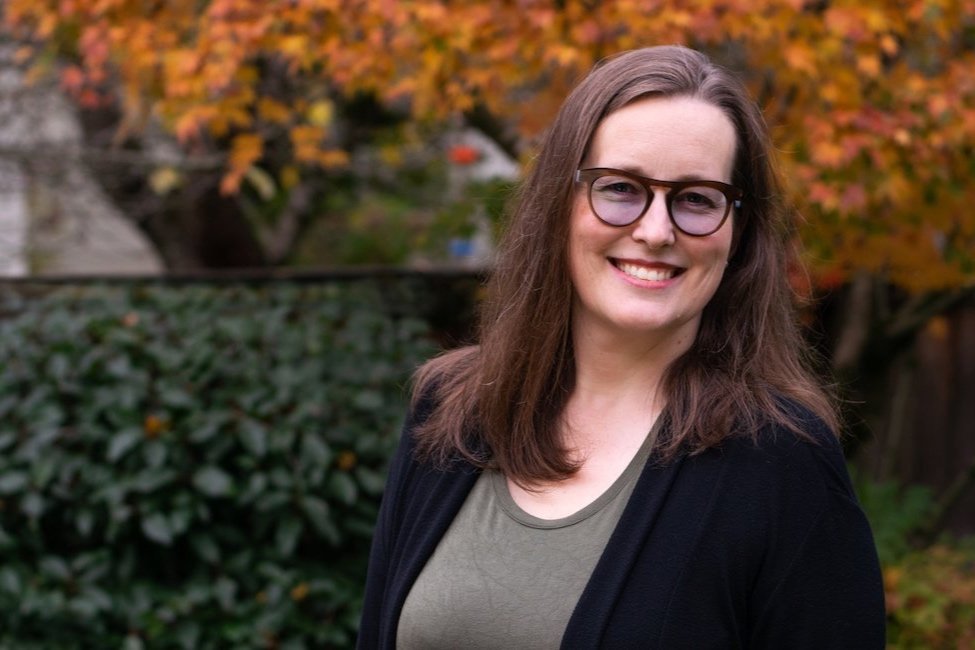 A picture of Melissa Taylor, smiling towards the camera, standing infront of some trees and plants.